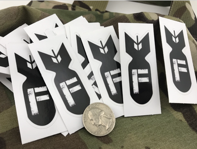  A collection of F Bomb stickers spread over camo fabric, with a quarter for size.